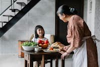 Delighted little Asian girl smiling and speaking with grandmother while having lunch at home