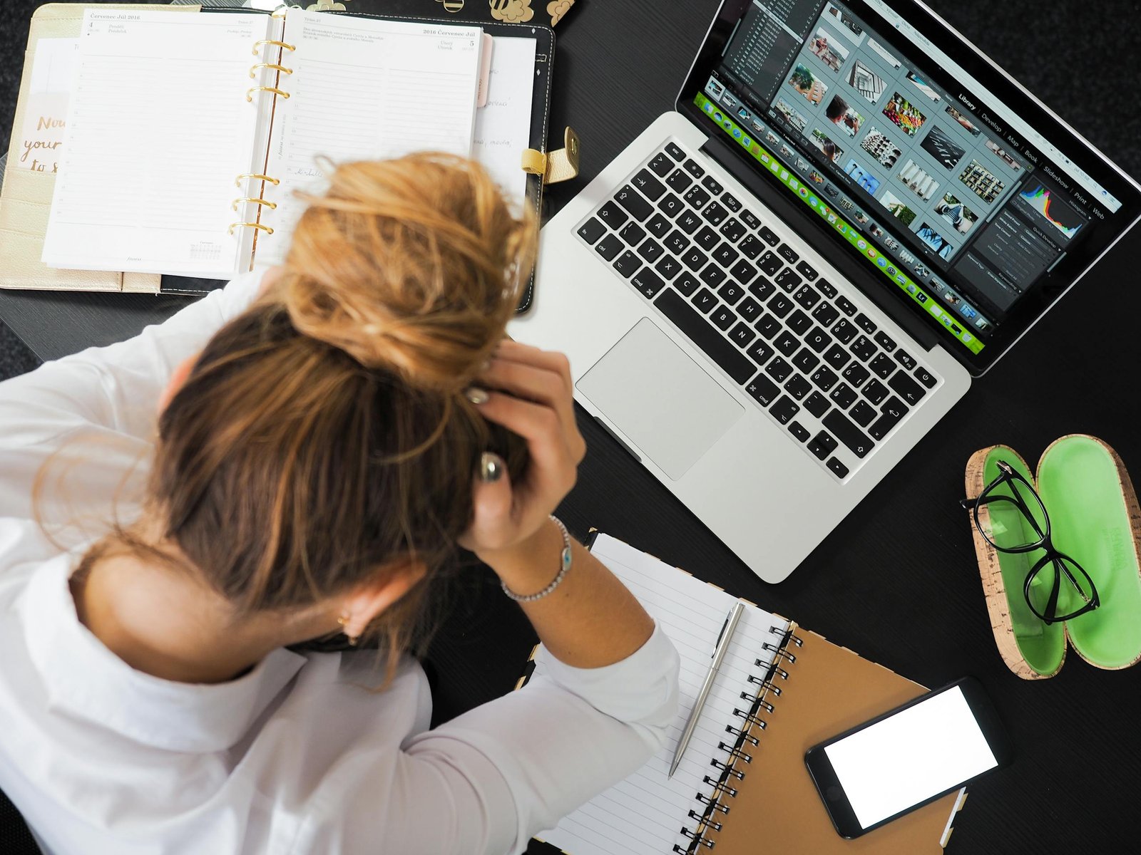 Overhead view of a stressed woman holding her head while working at a desk with a laptop, phone, and notebooks — concept for how to stop overthinking and mental overload.