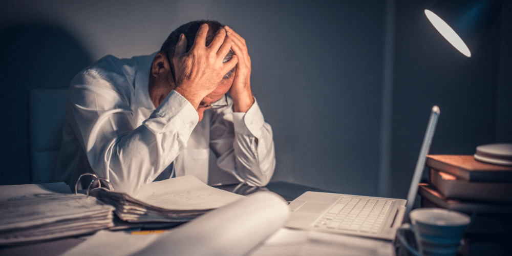 Tired employee sitting at desk with hands on head in a dimly lit office, illustrating workplace burnout and stress.
