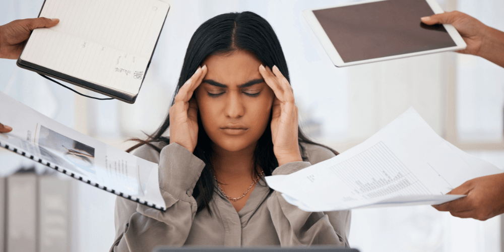 A stressed woman surrounded by papers and electronic devices, illustrating work pressure and burnout.