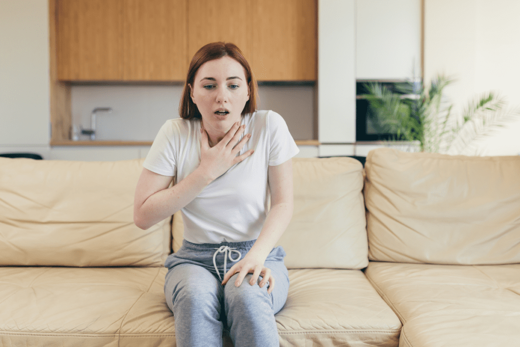 A young woman sitting on a couch, holding her chest with a distressed expression, indicating anxiety or emotional overwhelm.