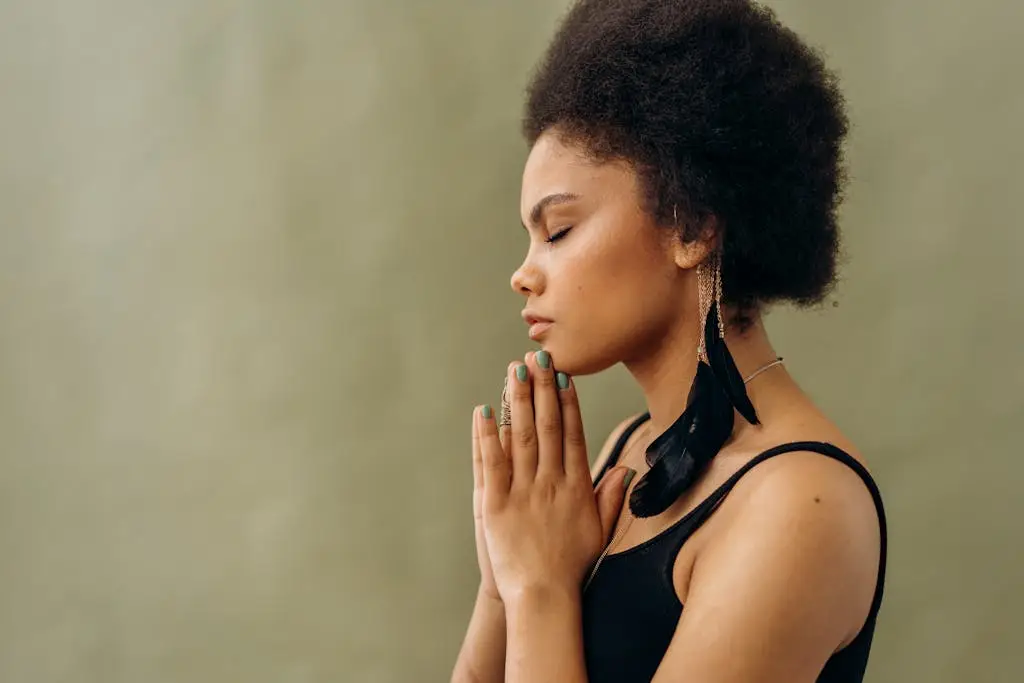 A young woman with afro hair practicing calming meditation indoors, eyes closed and hands together in a peaceful posture.