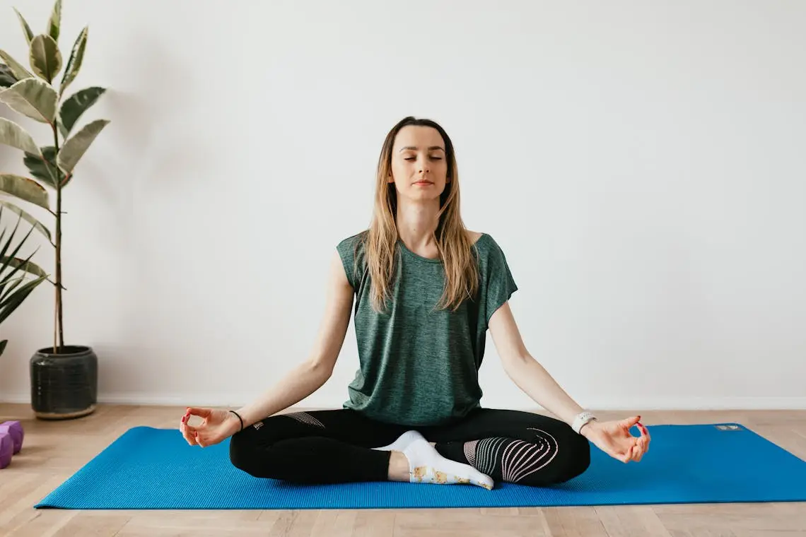 A serene blonde woman meditating on a yoga mat at home, sitting cross-legged with eyes closed near a potted plant, radiating calm and balance.