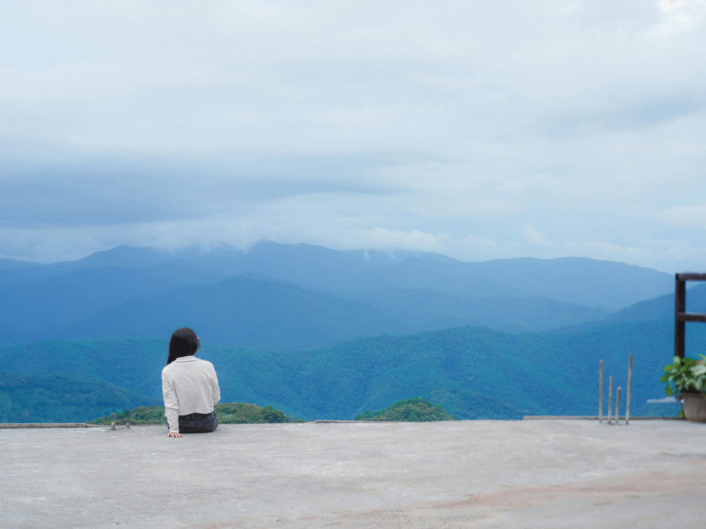A woman sitting alone facing mountain views, embodying solitude and creativity in a peaceful natural setting.